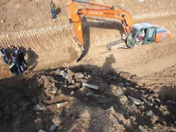 墓顶停放着一辆橙黄色的大型挖掘机,土层有明显的挖掘痕迹。